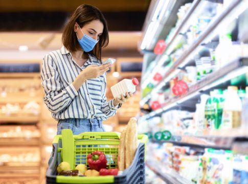 Young woman in disposable face mask taking dairy products from shelf in the supermarket, holding bottle and smartphone, scanning bar code on product through mobile phone, walking with trolley cart