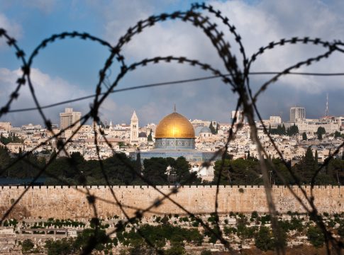 The Old City of Jerusalem, including the Dome of the Rock and various church steeples, seen through coils of razor wire, illustrating the Holy Land's history of division and conflict.