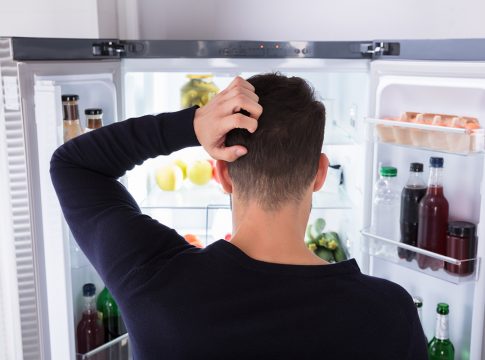 Rear View Of A Confused Young Man Looking At Food In Refrigerator